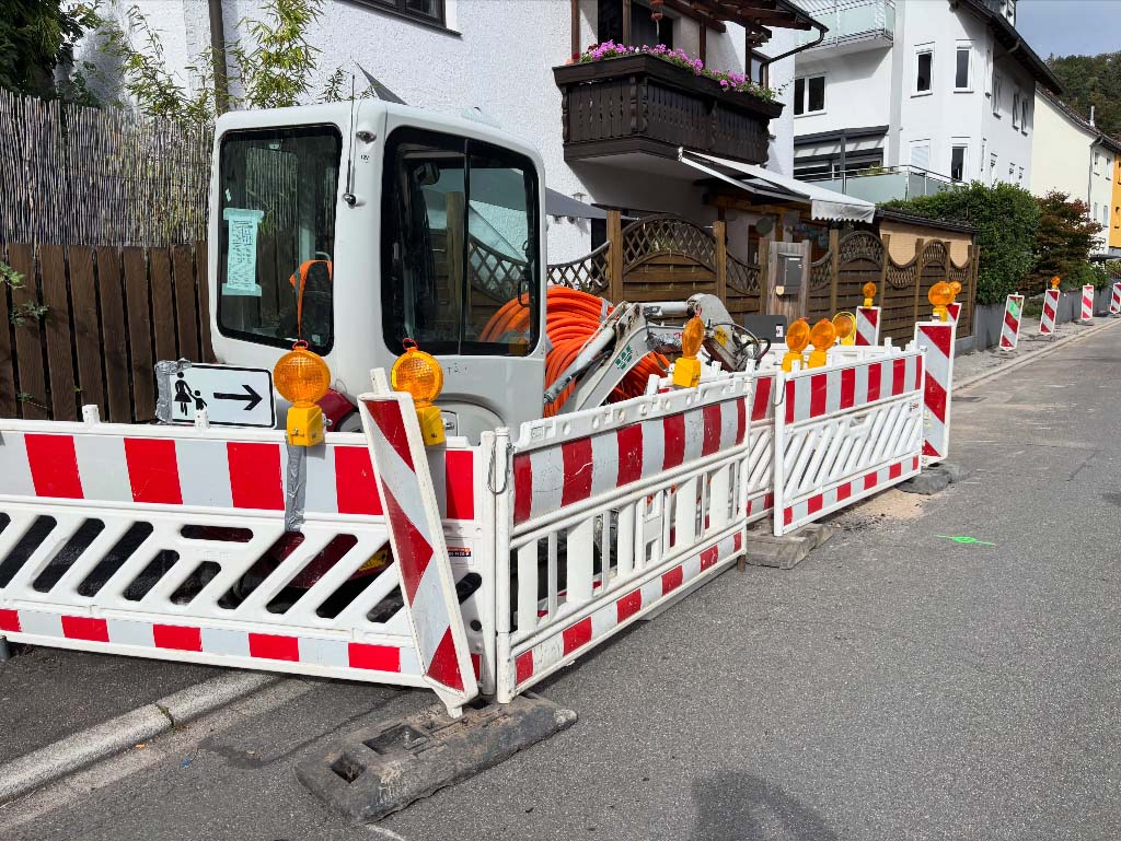 Bagger hinter rot-weißen Absperrgittern, daneben eine orange Glasfasertrommel vor Wohnhäusern in einer Gaiberger Straße
